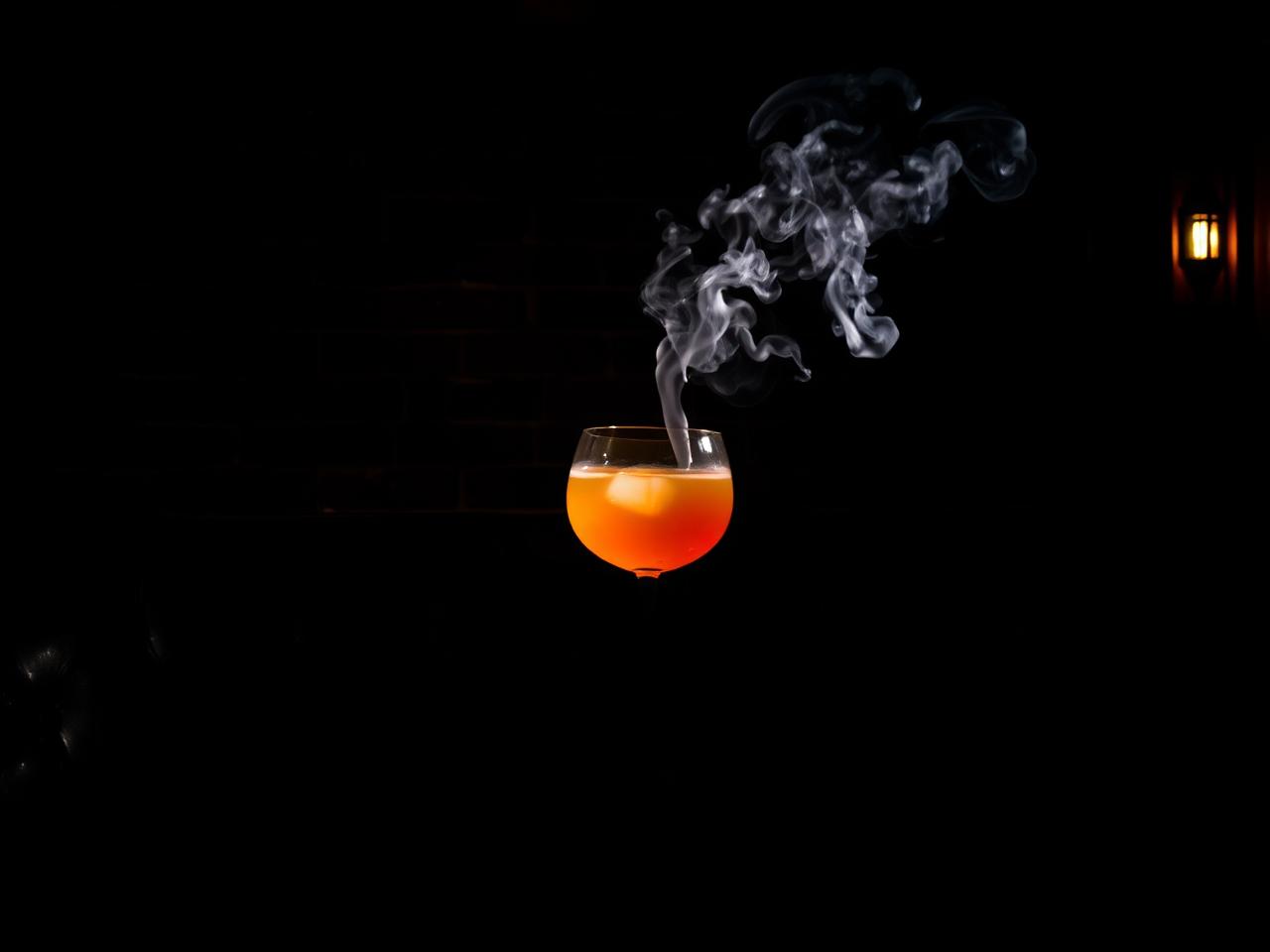 Intimate Manchester speakeasy cocktail bar with dark moody lighting and a single beautifully crafted smoking cocktail in a coupe glass against a near black background